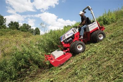 Person operating a red mower on a grassy hillside under a blue sky with clouds.