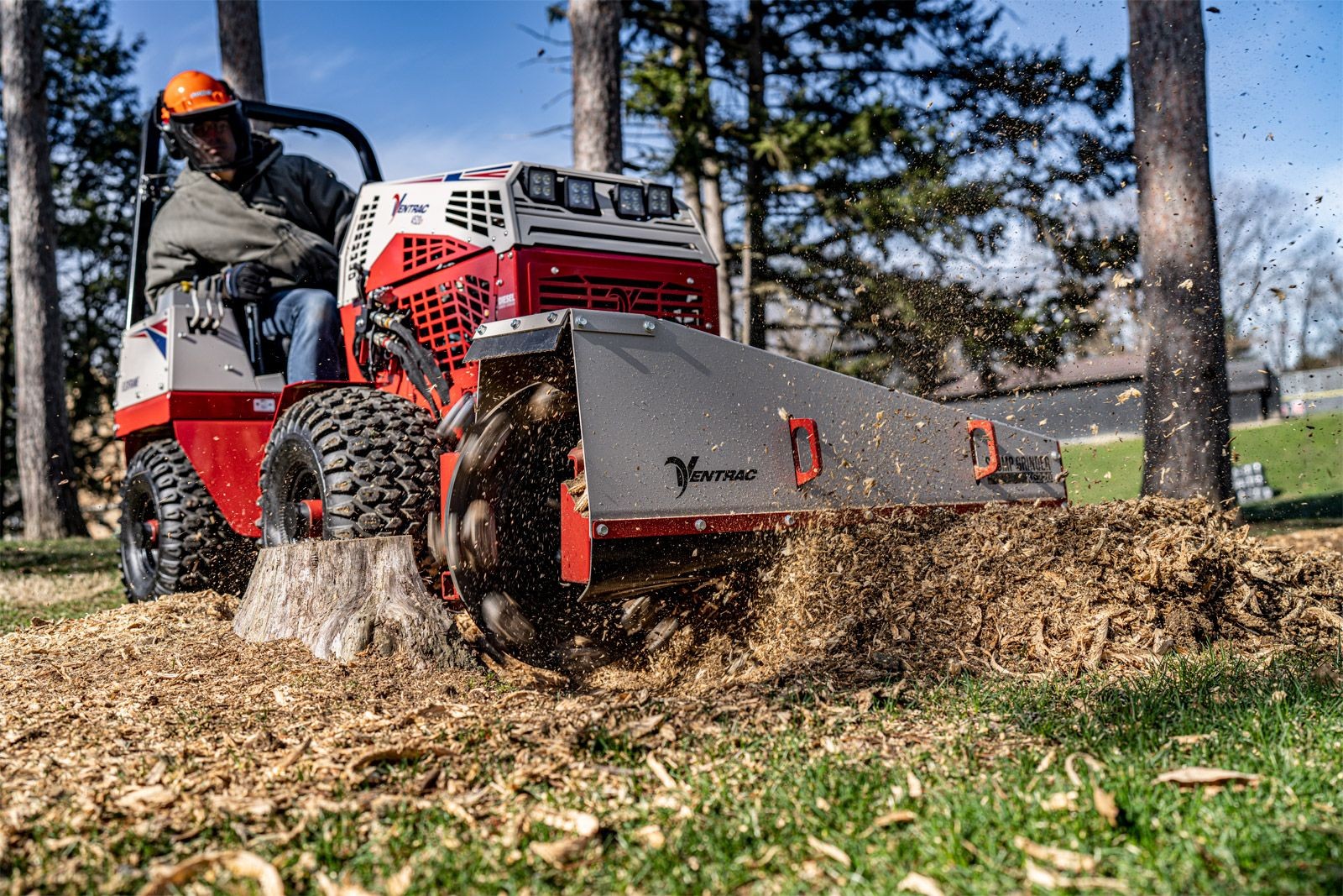 Person operating red stump grinder on grassy field, wood chips flying.