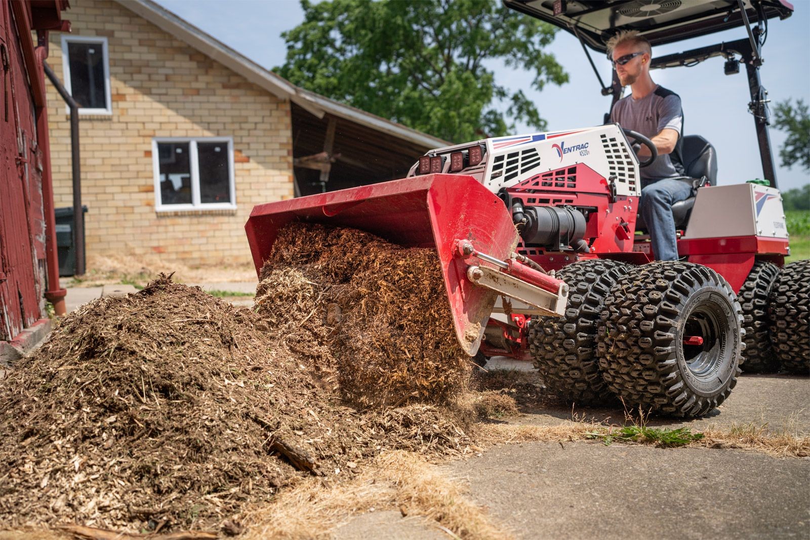 Person operating a large red landscaping machine, unloading mulch onto the ground near a building.